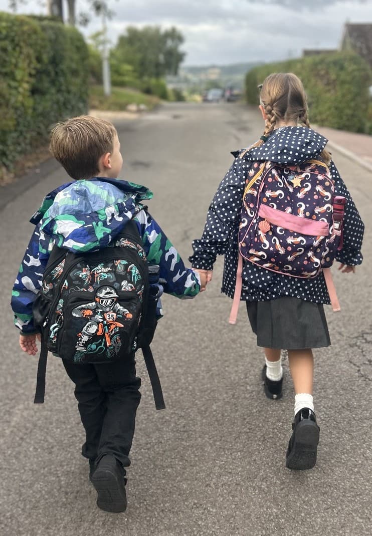 Two children in school uniform walking up a lane hand in hand, seen from behind with their backpacks on