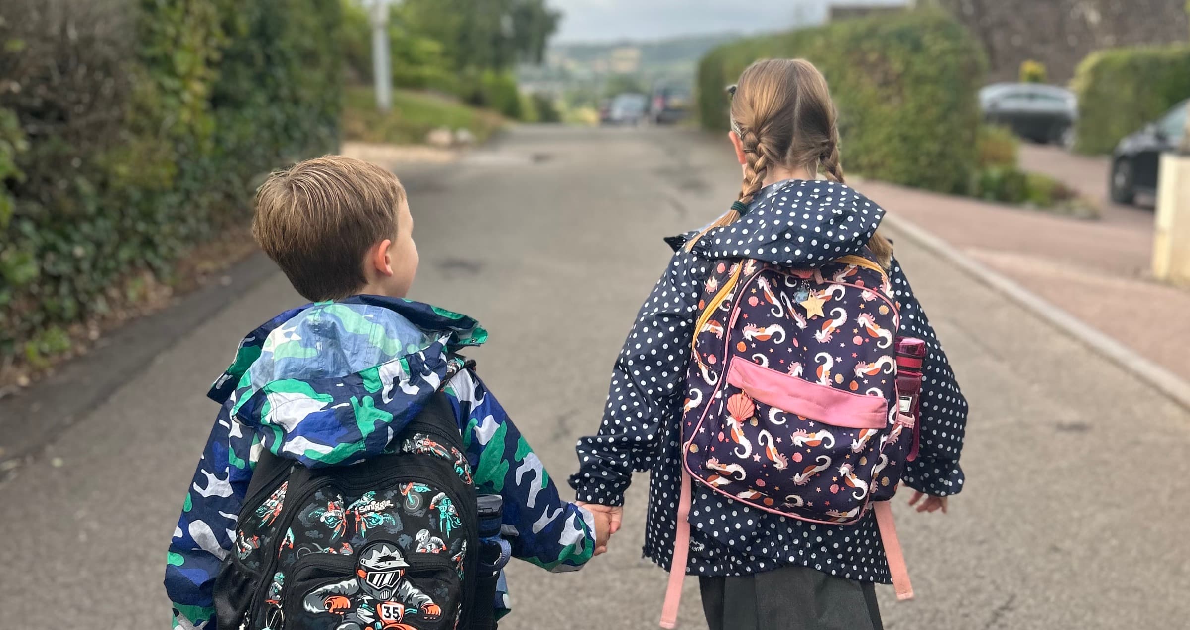 Two children in school uniform walking up a lane hand in hand, seen from behind with their backpacks on