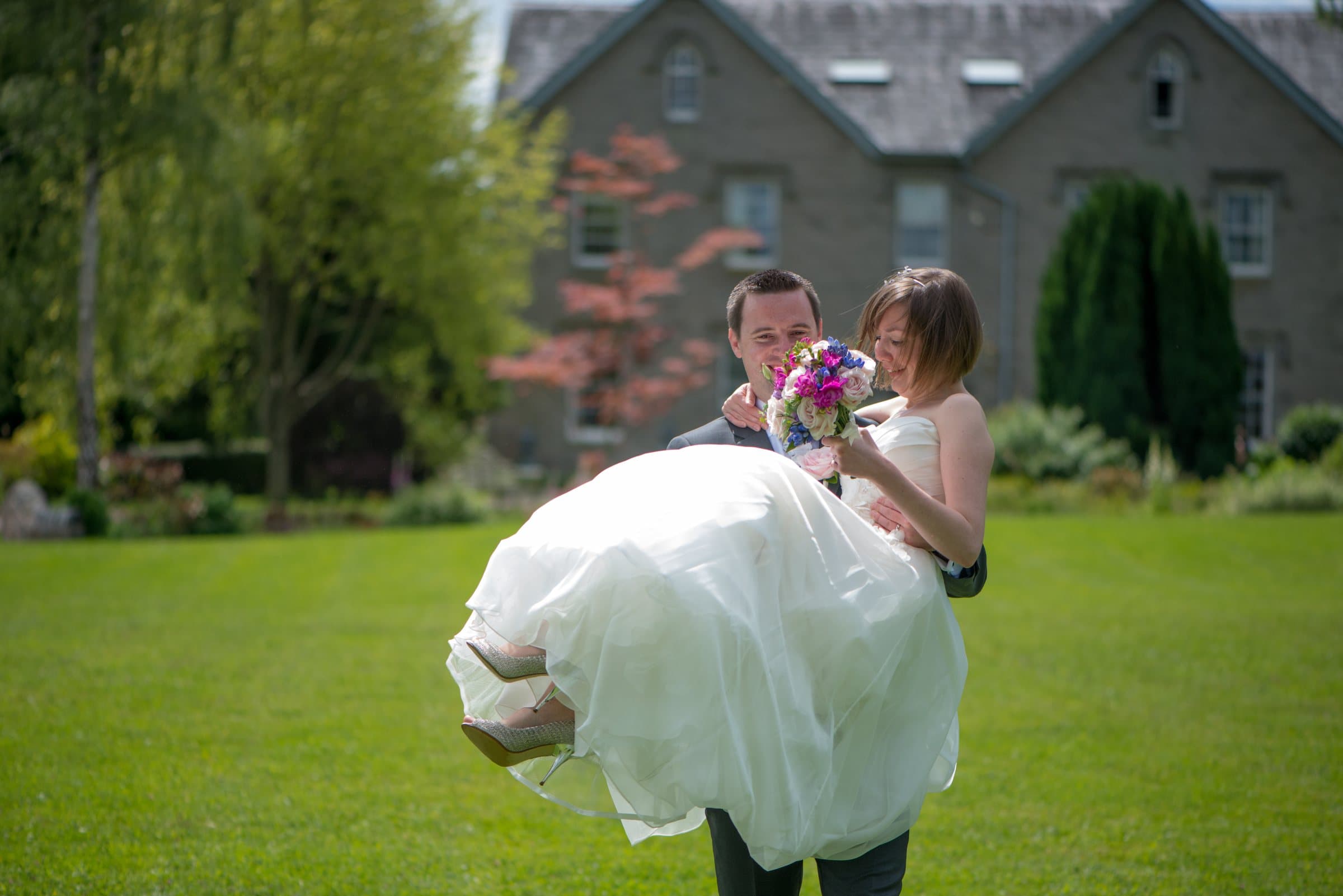 A groom carrying his bride across the lawn on their wedding day